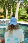 A person with curly hair wears a light blue Charleston Pineapple Embroidered Hat and a T-shirt with a coastal design, relaxing poolside amid lush trees.