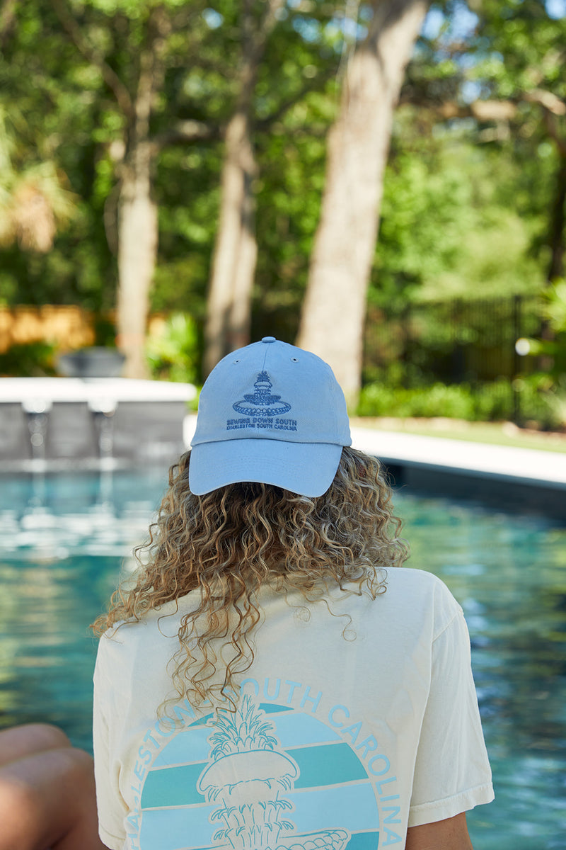 A person with curly hair wears a light blue Charleston Pineapple Embroidered Hat and a T-shirt with a coastal design, relaxing poolside amid lush trees.