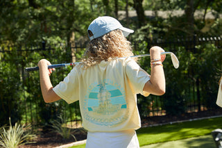 A person with curly hair and a Charleston Pineapple Embroidered Hat holds a golf club behind their back. They wear a yellow T-shirt featuring a graphic and text on the back. Trees and a fence in the background add charm to this relaxed outdoor scene.