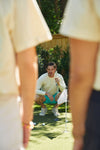 On a mini-golf course, someone crouches with a putter, focused on the ball, surrounded by trees and a wooden fence. In the foreground, two others partially visible are wearing Charleston Pineapple Crewnecks, adding a playful vibe to their game.