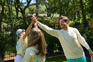 Three people wearing matching Charleston Pineapple Crewneck Sweatshirts high five and smile outdoors in a sunny, green garden with trees and a black metal fence in the background.