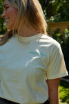 A woman beams under the sun, wearing the Charleston Pineapple Tee with SEWING DOWN SOUTH printed on it. Her blonde hair flows over the shirt, complementing the lush greenery behind her—capturing Charlestons iconic pineapple fountain charm perfectly.