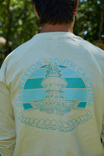 Wearing a Charleston Pineapple Crewneck with a green pineapple fountain graphic and Charleston, South Carolina text on the back, someone stands outdoors at Waterfront Park amidst blurred trees in a serene moment.