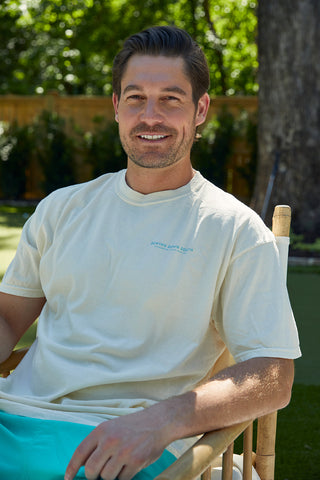 A bearded, short-haired individual smiles while sitting outdoors in a Charleston Pineapple Tee and turquoise shorts, surrounded by lush greenery and a wooden fence, enjoying the serene ambiance of a sunny Charleston day.
