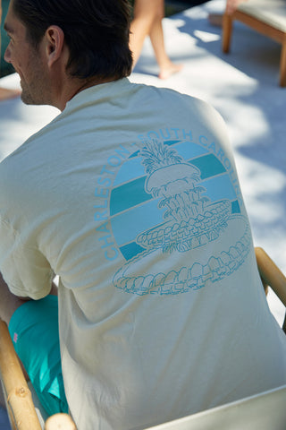 A man sits on a wooden chair wearing the Charleston Pineapple Tee, a light beige shirt featuring a blue-green pineapple fountain design and Charleston, South Carolina on the back. The sun casts playful shadows on the wooden floor.