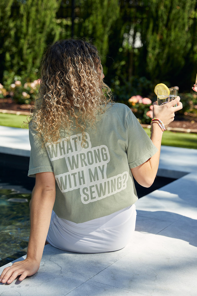 A person with curly hair lounges by the pool, holding a lemon drink and wearing a light green WWWMS Short Sleeve Tee, Sandstone, made from 100% ring spun cotton. The tee has WHATS WRONG WITH MY SEWING? on the back. Trees and vibrant flowers frame the scene.