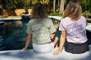 Two women sit poolside in WWWMS Short Sleeve Tees, Sandstone, featuring Whats wrong with my sewing? on the back. The unisex fit and 100% ring-spun cotton promise comfort, as they enjoy drinks under trees beside a fence.
