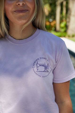 A person wears an Orchid WWWMS Short Sleeve Tee with a vintage sewing machine design, made from 100% ring spun cotton. The text reads SEWING DOWN SOUTH, EST 2019, CHARLESTON, SOUTH CAROLINA, set against an outdoor backdrop with trees.