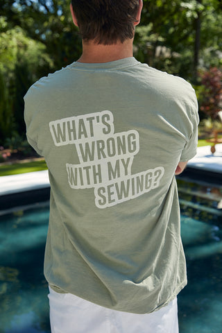 By a pool surrounded by lush greenery, someone wears the comfy WWWMS Short Sleeve Tee in Sandstone, featuring a unisex fit made of 100% ring-spun cotton. Bold white letters on the back question, WHATS WRONG WITH MY SEWING?.