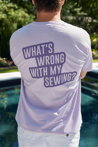 A person with short hair stands by a pool, wearing the WWWMS Short Sleeve Tee in orchid. The unisex fit shirt, made from 100% ring spun cotton, features WHATS WRONG WITH MY SEWING? on the back. It beautifully contrasts with the lush greenery and sparkling water nearby.