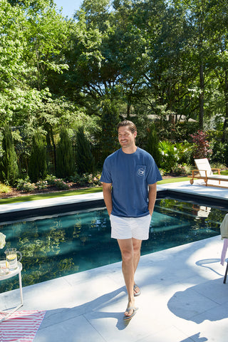 Someone in an SDS Vintage Logo Pocket Tee and white shorts strolls by a pool with greenery. Nearby, a lounge chair and small table with drinks enhance the sunny, relaxed vibe.