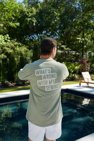 A person in a lush garden by a pool wears the Whats Wrong With My Sewing? Long Sleeve Tee in sandstone, featuring an unisex fit and bold white text on the back.