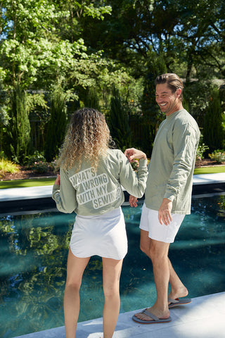 A woman with curly hair and a man are by a pool, both in Whats Wrong With My Sewing? long-sleeve tees in sandstone. Hers is paired with a white skirt, while his matches with white shorts. They hold hands and smile warmly.