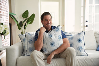 A man in a blue shirt and beige pants sits on a white sofa, smiling with the Coastal Collections Navy Starburst Pillow. The bright room, decorated with plants and a brick wall, enhances the stylish charm of Sewing Down South decor.