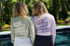 Two people with curly hair stand by a pool, facing away. Theyre wearing Whats Wrong With My Sewing? Long Sleeve Tees in Orchid, unisex fit, 100% ring spun cotton, with the bold phrase printed on the back. The backdrop is lush with greenery.