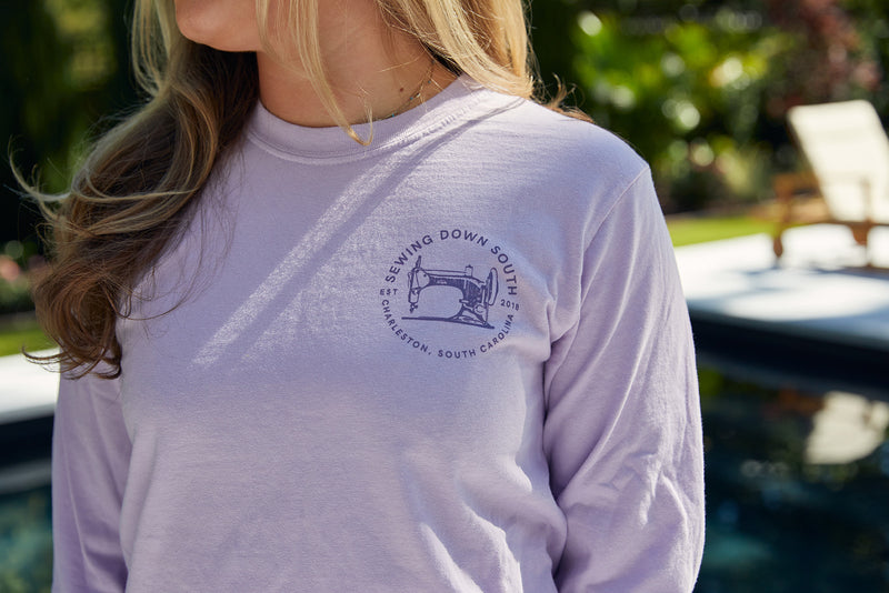 By the pool, someone wears the Whats Wrong With My Sewing? Long Sleeve Tee in Orchid, crafted from 100% ring-spun cotton. This unisex tee features a Sewing Down South logo with a sewing machine and Charleston, South Carolina, exuding comfort amid the green surroundings.