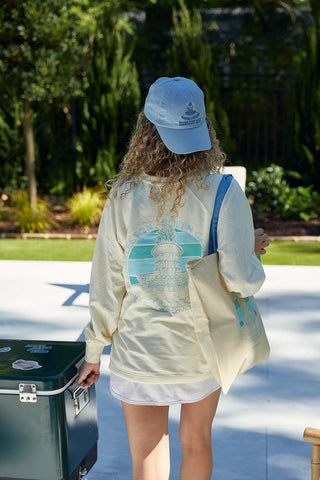 A person with curly hair, dressed in a cream Charleston Pineapple Crewneck Sweatshirt and a light blue cap, stands outdoors with a tote bag. A cooler and greenery are visible in the background.