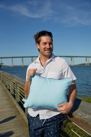 A smiling man in a white shirt stands on a wooden boardwalk by the water, holding the Dockside Knots Lumbar Outdoor Pillow. A bridge and clear blue sky appear in the background.