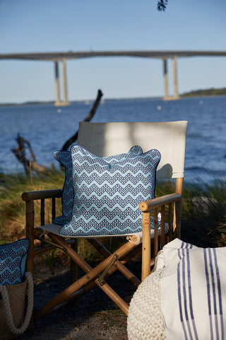 A wooden chair with a Dockside Chevron Ropes Outdoor Pillow sits by the water’s edge, with a bridge in the background and a blanket and basket nearby, creating a relaxed outdoor setting.