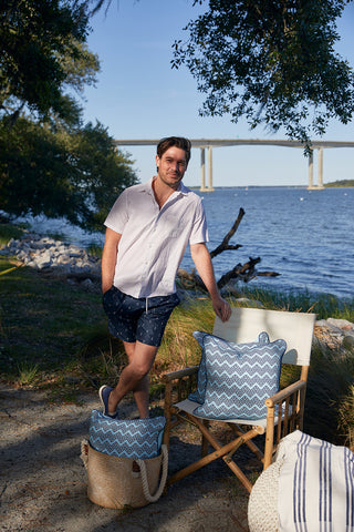 A man in a white short-sleeve shirt and patterned shorts stands by a wooden chair with Dockside Chevron Ropes Outdoor Pillow, near a riverbank with a bridge in the background and shade from nearby trees.