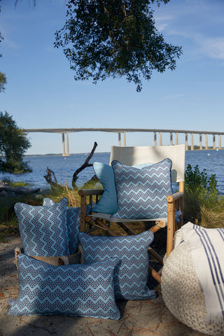 A group of Dockside Chevron Ropes Outdoor Pillows with blue patterns rest on the ground and a wooden chair by a riverbank under blue skies, with trees and a bridge in view for a relaxing outdoor scene.
