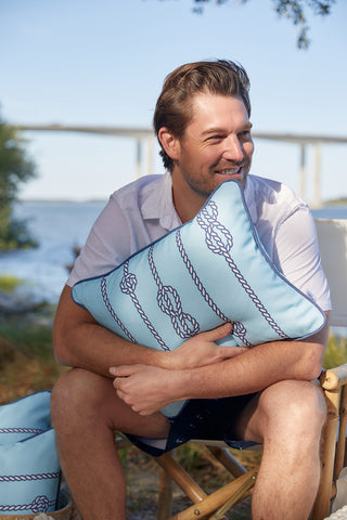 A man smiles by the water, holding a light blue Dockside Knots Lumbar Outdoor Pillow from the Coastal Collection. Made with SDS Shield® Outdoor Fabric and featuring a rope knot pattern, it’s set against a backdrop of trees and a bridge.