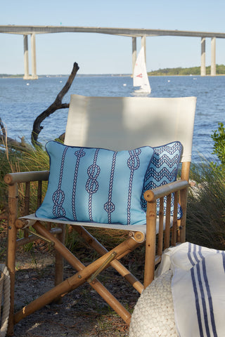 A bamboo chair with a white outdoor fabric seat and the Dockside Knots Lumbar Outdoor Pillow sits by the water. A bridge and sailboat are visible in the background, while grass and a striped blanket appear in the foreground.