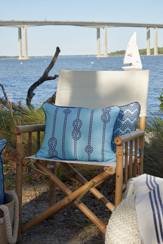 A bamboo chair with a Dockside Knots Lumbar Outdoor Pillow sits by the water, grasses and a bridge behind. A white sailboat glides by in the sun, showcasing the charm of the Sewing Down South™ pillow pattern.