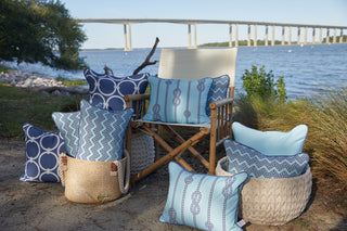 A wooden chair by the riverside is surrounded by blue-patterned pillows from the Coastal Collection, including the Dockside Knots Lumbar Outdoor Pillow in woven baskets, set on sandy ground with grass, trees, and a bridge nearby.
