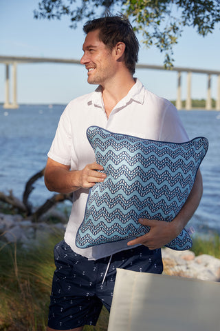 A man in a white shirt and navy anchor-print shorts stands by the water, smiling while holding a Dockside Chevron Ropes Outdoor Pillow. A bridge and trees are visible in the background.