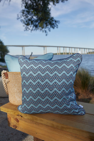 The Dockside Chevron Ropes Outdoor Pillow in blue and white rests on a wooden bench by the water, with a woven basket, another pillow, and a bridge with trees in the distance beneath a clear blue sky.