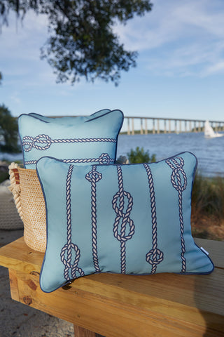 Two Dockside Knots Lumbar Outdoor Pillows with rope-knot designs are set on a wooden bench outside. Behind them, water, a bridge, trees, and a sailboat evoke the coastal charm of this accent from the Coastal Collection.