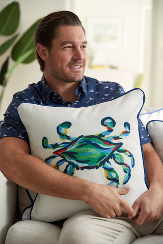 A man relaxes on a couch, clutching The King Crab Pillow with Navy. His blue shirt, featuring small boat patterns, complements the vibrant design of this Sewing Down South item. A lush green plant adds a touch of nature to the scene.