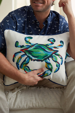 A person in a blue patterned shirt holds The King Crab Lumbar Pillow with Navy from the Coastal Collection, displaying vibrant hues of green, blue, and red. The image captures their relaxed demeanor, though they are partially cropped out.