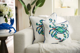 The King Crab Lumbar Pillow with Navy from the Coastal Collection adds charm to the white sofa, while a nearby table displays a bouquet of white flowers and a blue crab figurine. A large leafy plant in the background completes this Sewing Down South scene.