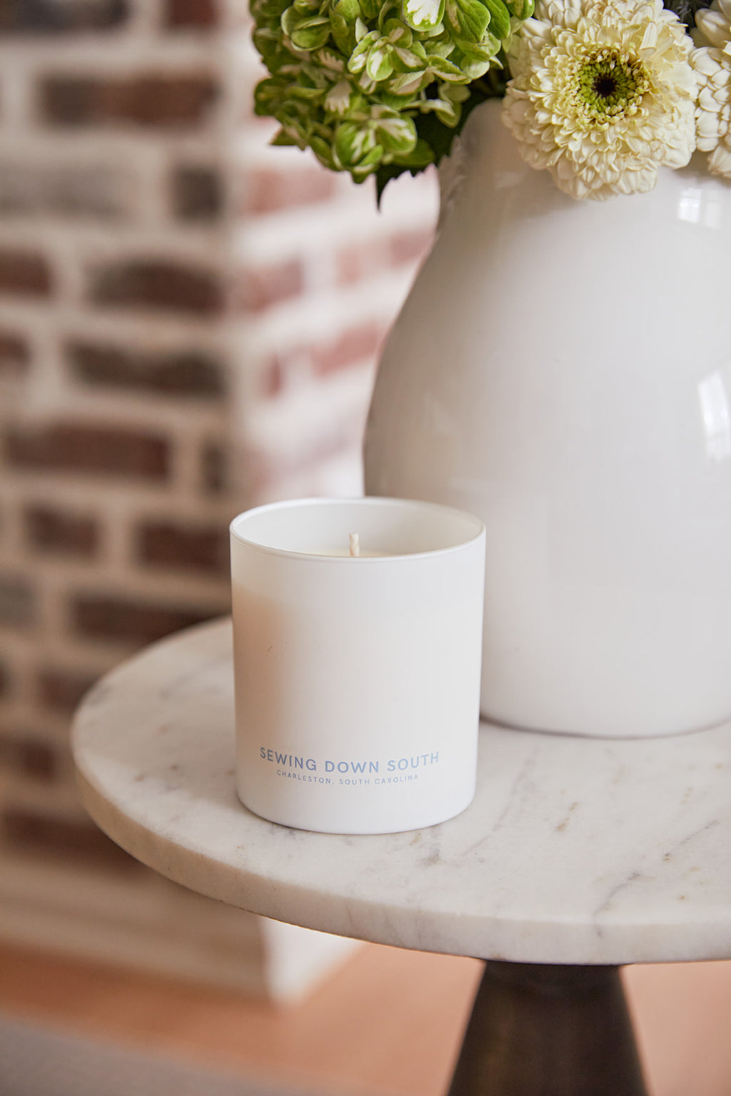 A white soy candle labeled No. 2 - Cobblestone Flower Candle sits on a round marble table. Behind it, a white vase holds green hydrangeas and white flowers, while the rustic charm of a brick wall completes the serene backdrop with hints of honey berries musk lingering in the air.