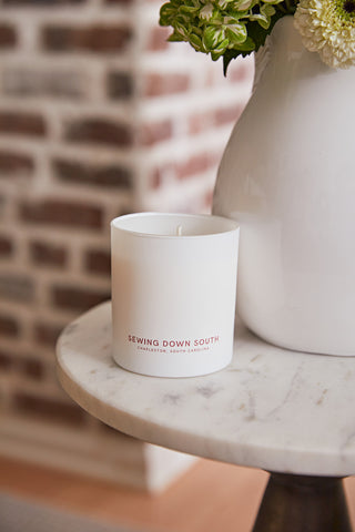A No. 4 - Cotton Blossom Candle rests elegantly on a marble table beside a white vase of fresh flowers. A brick wall backdrop enhances the rustic charm and serene setting, with Sewing Down South, Charleston, South Carolina prominently on the candle.