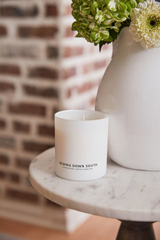 A white No. 1 - Charleston Proper Candle rests on a round marble table, releasing a strong fragrance. Beside it, a white vase holds green and white flowers against a charming brick wall backdrop, capturing Charlestons essence.