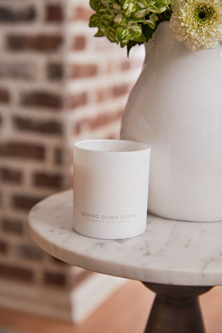A white No. 3 - Midnight Stroll Candle labeled Sewing Down South, Charleston, South Carolina sits on a round marble table, filling the air with eucalyptus and cedar scents. In front of a blurred brick wall backdrop is a white vase with green and white flowers.