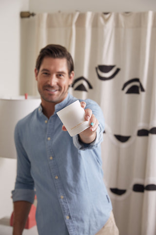 A man in a light blue shirt smiles and holds the Mystery Candle + Room Spray Bundle toward the camera. Behind him, there’s a curtain with black abstract shapes and a lamp.