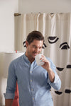 A man in a light blue button-up shirt enjoys his coffee near a lamp, smiling as the No. 5 - Sugar Cookie Candle fills the air with a vanilla scent. The cozy setting is complemented by the geometric black-patterned curtain behind him.