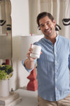 A smiling man in a light blue shirt presents the No. 1 - Charleston Proper Candle named SOUTHERN GENTLEMAN in a stylishly decorated room with a unique lamp and bouquet, enhancing the cozy atmosphere with its high fragrance percentage soy formula.