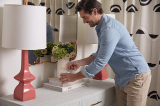 Wearing a light blue shirt, a man lights a No. 4 - Cotton Blossom Candle on the dresser. Surrounded by fresh florals, stacked books, and two pink lamps, his touch illuminates the room. A mirror and patterned curtain add to the tranquil backdrop.
