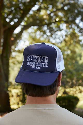 A man stands outdoors with softly blurred trees behind him, wearing a SDS Collegiate Trucker hat in navy and white. The hat features an embroidered SDS logo and Sewing Down South Est. 2018 on the front.