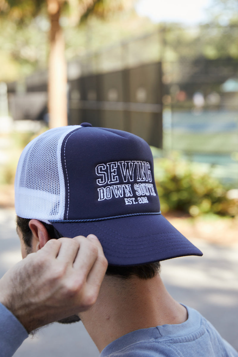 A person wearing the SDS Collegiate Trucker hat, in navy and white with the embroidered SDS logo and SEWING DOWN SOUTH EST. 2018 on the front, adjusts it while standing outdoors against a backdrop of blurred trees and a fence.