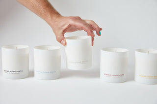 A hand with painted nails holds one of five white minimalist jar candles, labeled No. 1 - Charleston Proper Candle from Sewing Down South. These high-fragrance candles are elegantly arranged on a pristine white surface against a matching backdrop.