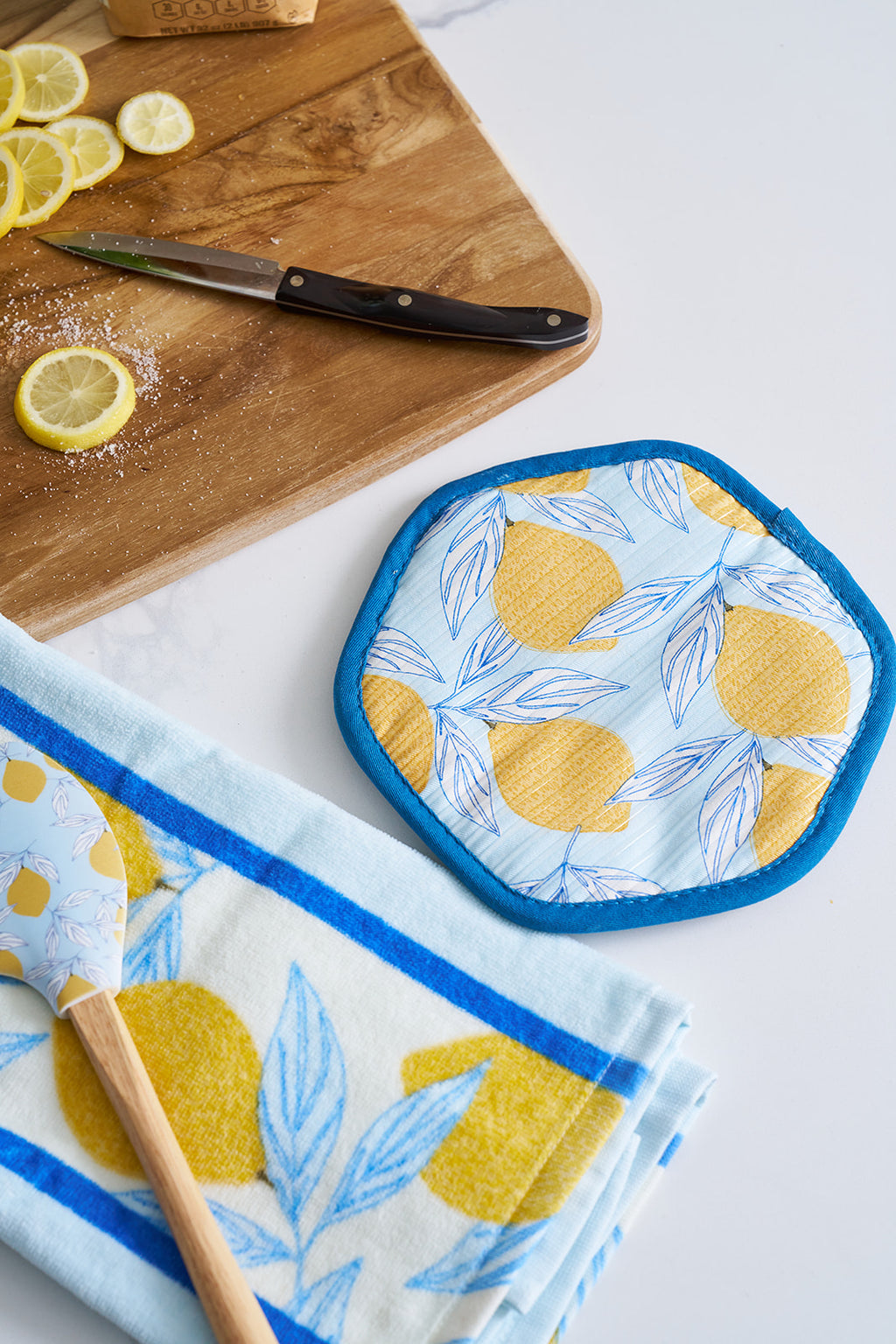 A cutting board with sliced lemons and a knife rests next to the Lemon Kitchen Bundle: a blue and yellow 100% cotton lemon-patterned kitchen towel set, pot holder, and spatula on a white countertop.
