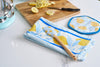 The Lemon Kitchen Bundle—featuring a lemon-patterned towel, spatula, and round pot holder—sits on a white counter. Sliced lemons and a knife are visible in the background on a wooden cutting board.