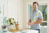 A person in a kitchen, wearing a patterned shirt, smiles holding a lemon. A Lots of Lemons Pot Holder is draped over their shoulder. The counter displays a mixer, sugar bag, cutting board with lemon slices, and flowers in a vase.