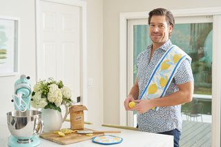 A man stands in a bright kitchen with the Lemon Kitchen Bundle—featuring a lemon-patterned kitchen towel, pot holder, and spatula—over his shoulder, next to a stand mixer, flowers, brown sugar, and sliced lemons.